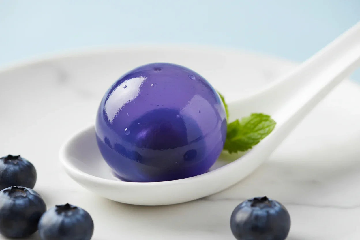Popball bleu violet brillant posé sur une cuillère blanche avec des myrtilles et une feuille de menthe